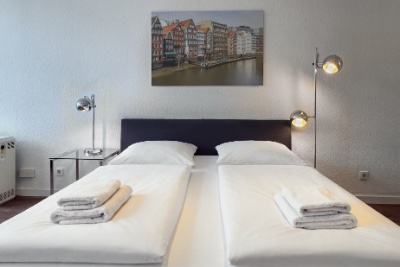 Doppelzimmer Fleetblick: Doppelbett mit dunklem Kopfteil, gestapelten Handtüchern, Glasnachttisch und Stehlampe unter einem Wandbild