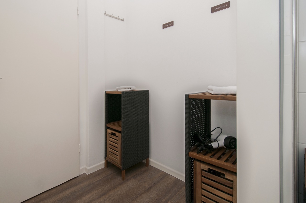 Doppelzimmer Fleetblick: Flurbereich mit zwei Holzregalen mit Handtüchern und Föhn, Wandhaken und Schildern an weißer Wand