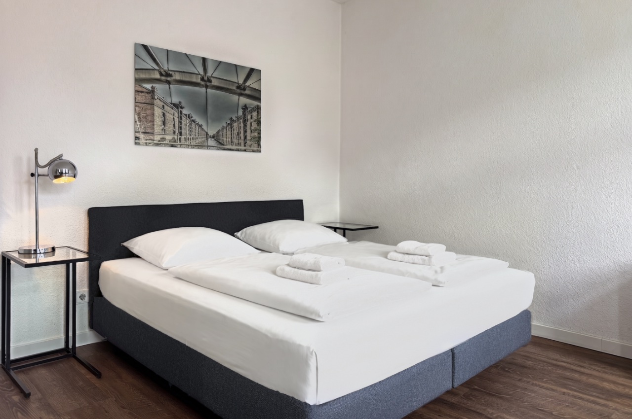 Habitación doble Fleetblick: Cama doble con ropa blanca y toallas, mesita con lámpara de lectura y cuadro en la pared