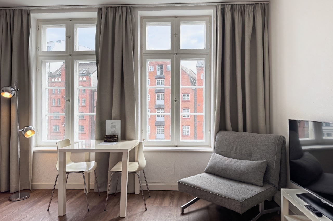 Habitación doble Fleetblick: Zona de estar con mesa blanca, dos sillas, sillón gris, televisor y dos ventanas con cortinas
